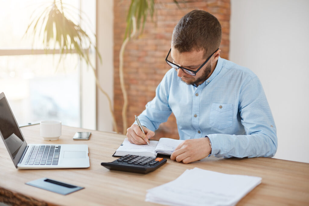 Business Concept. Young Unshaved Caucasian Male Manager In Glasses Sitting In Company Office, Writing Down Dates Of Meeting With Customers In Notebook With Concentrated Expression. - Abrir Empresa Simples