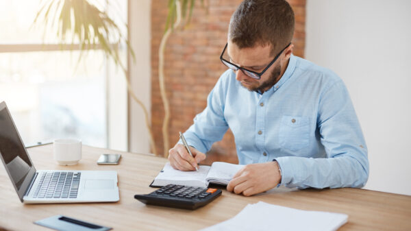 Business Concept. Young Unshaved Caucasian Male Manager In Glasses Sitting In Company Office, Writing Down Dates Of Meeting With Customers In Notebook With Concentrated Expression. - Abrir Empresa Simples