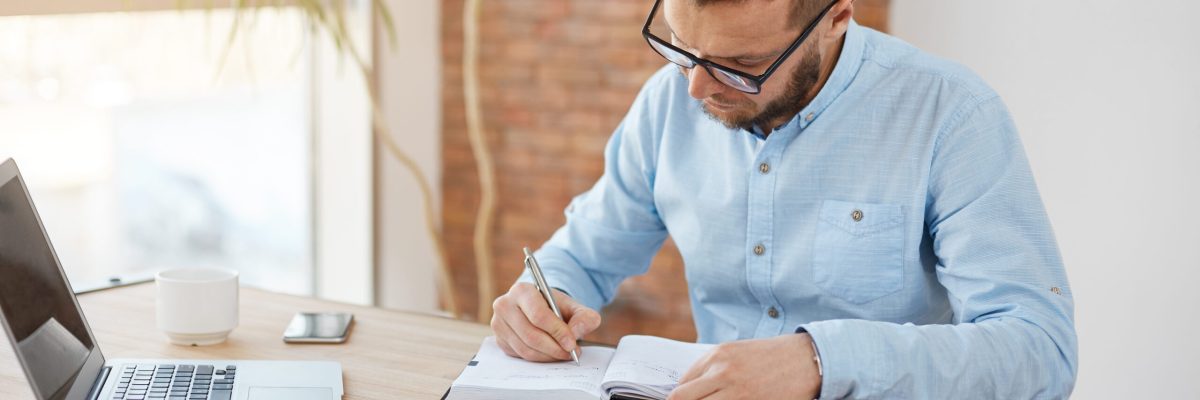 Business Concept. Young Unshaved Caucasian Male Manager In Glasses Sitting In Company Office, Writing Down Dates Of Meeting With Customers In Notebook With Concentrated Expression. - Abrir Empresa Simples